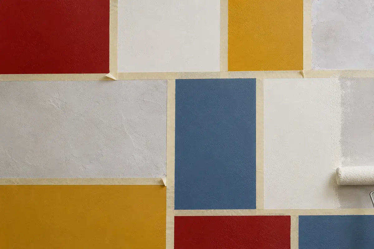 A wall divided by masking tape into blocks of red, ochre, blue and off-white paint, with a roller at the edge